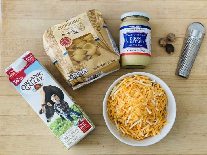 In the photo are the five main ingredients for the base recipe of No-Boil Overnight Mac & Cheese. They include milk, pasta shells, Dijon mustard, nutmeg and grated cheese. The recipe is easy to customize with your favorite combinations of cheese and seasonings.
