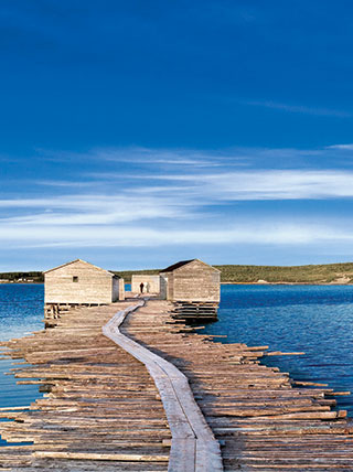 A winding wooden boardwalk leads to two weathered fishing stages on a pier in the blue waters of Newfoundland, Canada.