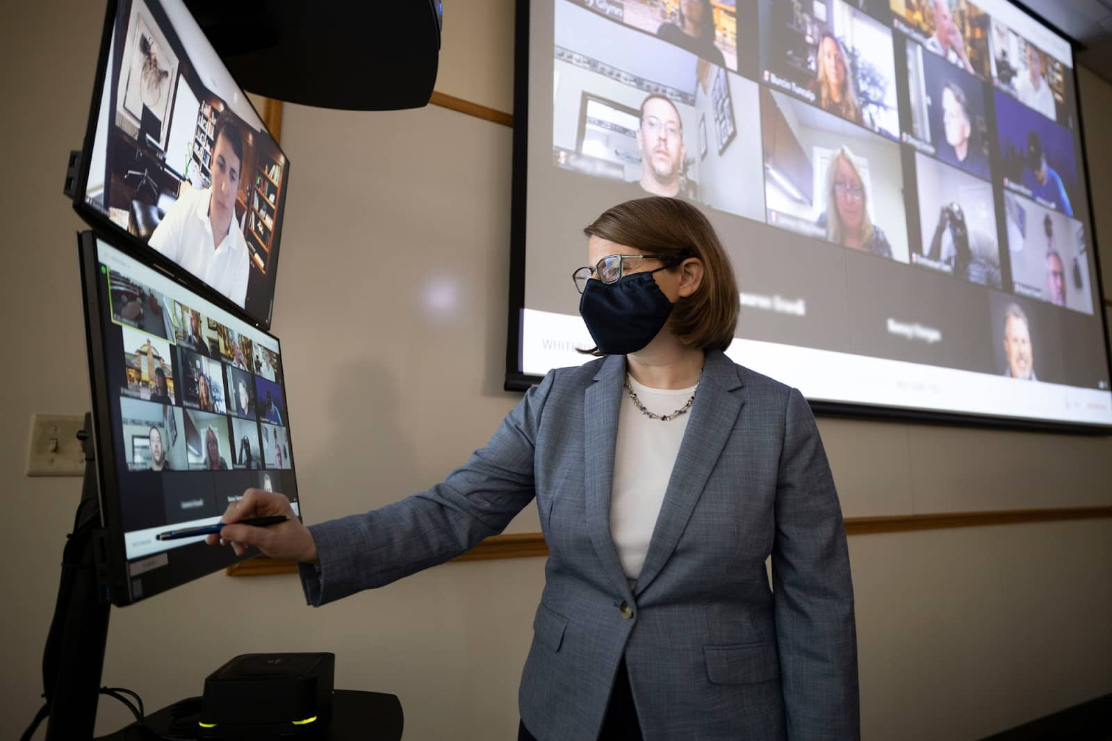 Woman wearing mask standing in front of dual monitors with Zoom video gallery of students, and large gallery wall of video meeting participants behind her